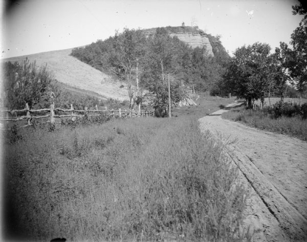 View down roadside of a dirt road, with a wooden fence, and hillside and a rock formation in the distance. Identified as Nicholas Park, located on the south side of Old Highway 54.