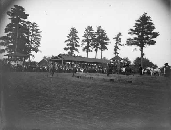 Outdoor view across racing track of a horse and racing buggy making a turn in front of a large grandstand. Probably at the Jackson County fairgrounds.