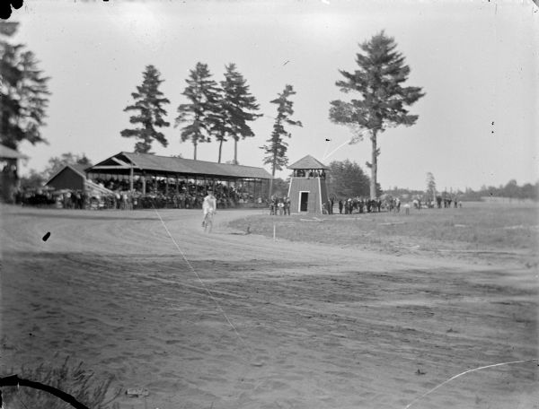 View across racing track towards a man on a bicycle turning a curve in front of a large grandstand. Probably at the Jackson County fair grounds.