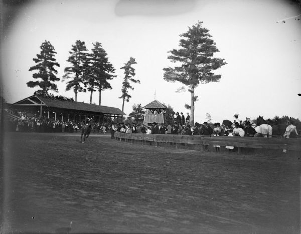 Outdoor view across tracks towards a man on a horse turning curve in a track in front of a large grandstand. Probably at the Jackson County fairgrounds.