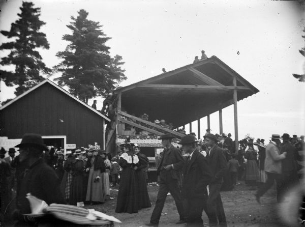 Outdoor view of people walking in front of a wooden grandstand. Probably at the Jackson County fairgrounds.