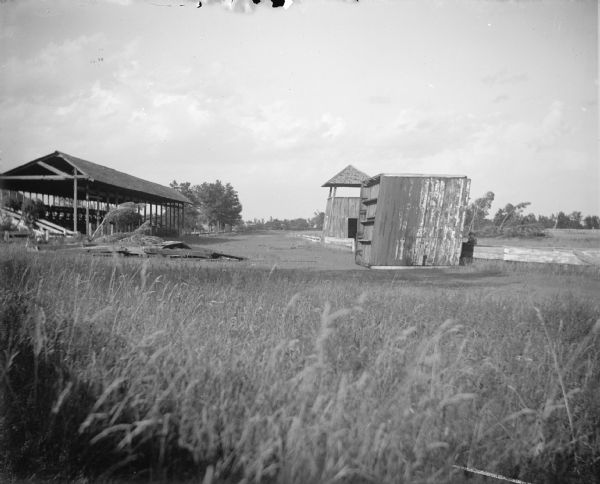 Outdoor view across grass towards a wooden structure and debris on the track between a wooden grandstand and viewing tower. Probably the Jackson County fairgrounds.