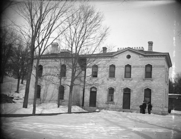 View across snow-covered ground towards a brick building. There are two men posing standing near the corner of the building on the right. The structure is identified as the Jackson County Jail and Sheriff residence located on Third Street behind the Jackson County Courthouse.