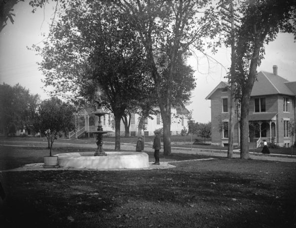 View across lawn towards two boys posing standing near a fountain. There is a church and a house along a sidewalk across the street. Identified as the fountain built on the lawn in front of the Jackson County Courthouse in 1906. The structure on the left is identified as probably the Lutheran Church, and the house on the right is owned by the Gebhardts.