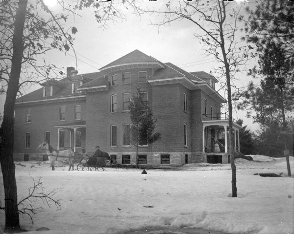 View across snowy ground towards a man posing sitting in a sleigh pulled by a single horse in front of a three-story brick building surrounded by snow. A woman is standing on the porch. Identified as the Jackson County Home located just south of the Jackson County fairgrounds, built in 1906 and torn down in 1974.