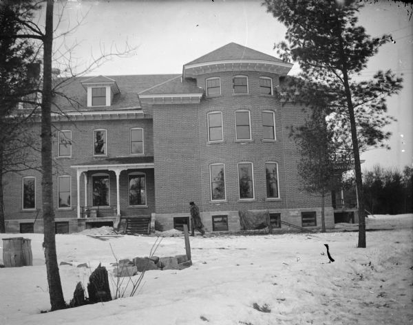 View across snowy ground towards a man walking in front of a three-story brick building surrounded by snow. Identified as the Jackson County Home located just south of the Jackson County fairgrounds, built in 1906 and torn down in 1974.