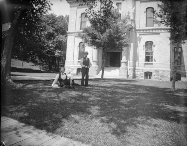 Outdoor portrait of two men, one posing sitting and one posing standing on the lawn in front of the Jackson County Courthouse, which was built in 1878 and located on the northwest corner of Main and Third Streets.