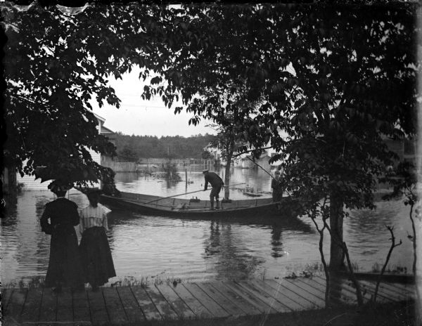 Outdoor portrait of two women posing standing on a wooden sidewalk near the water's edge. Behind them are two men in a long boat in the water among partially submerged buildings, probably during a flood.