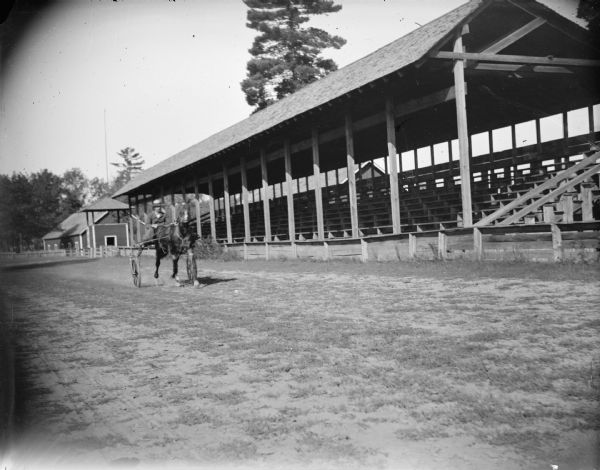 View towards a man posing sitting in a racing buggy pulled by a single horse in front of a wooden grandstand. Identified as the Jackson County fairgrounds.