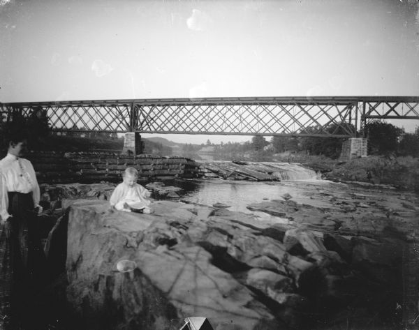 Outdoor view of a woman posing standing near a child posing sitting on large rocks. In the background is a railroad bridge over the Black River, and a river dam.