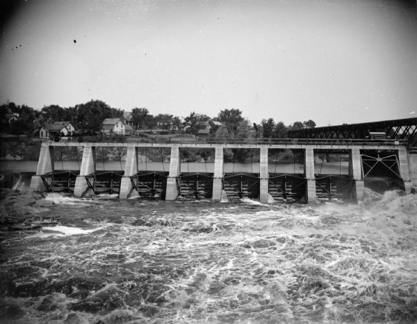 View upriver towards the dam in front, with the town of Black River Falls in the background on a hilly shoreline. There is a railroad bridge over the river on the far right. The gates for the dam were reportedly constructed in 1912.