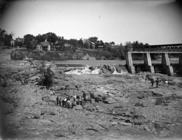 Outdoor view of a group of people on the rock in front of the dam located in front of Black River Falls, with several structures in the background. The gates for the dam were reportedly constructed in 1912.