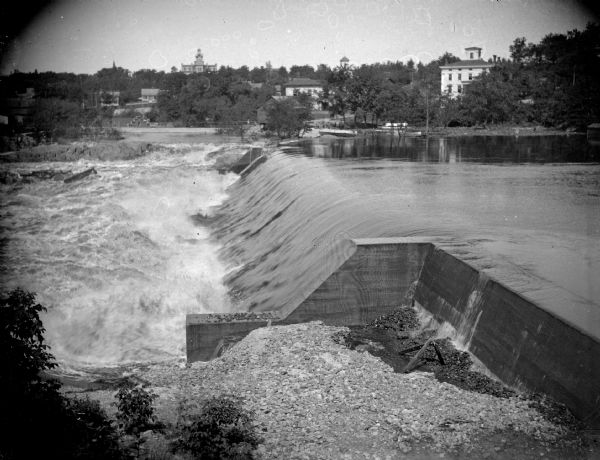 View from shoreline looking across the dam in front. Black River Falls is on the opposite shoreline, with several wooden structures, including the power house built in 1910. This photograph was taken between 1910 and the flood in October 1911.