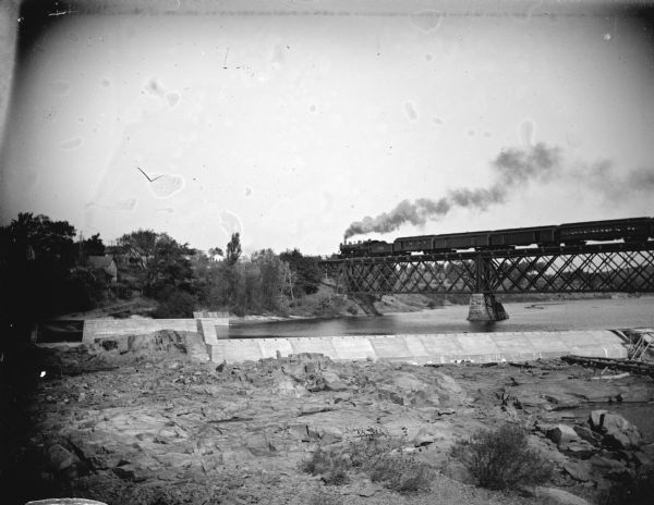 View of the dam in front of Black River Falls with several wooden structures in the background, and a train passing on the railroad bridge over the river.