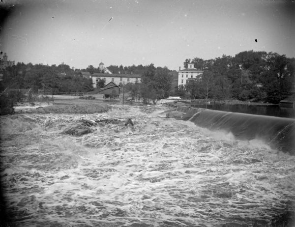 View from shoreline looking across the churning water just below the dam. On the opposite shoreline is the town of Black River Falls, with several wooden structures in the background, including the power house built in 1910. The photograph was taken between 1910 and the flood in October 1911.