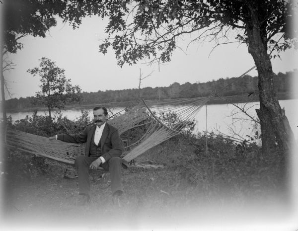 Outdoor portrait of a European American man posing sitting on a hammock with a body of water in the background. Man identified as Tom Van Schaick.