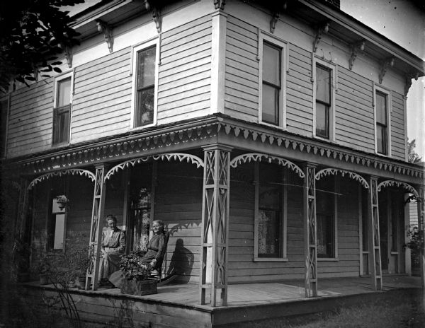 Outdoor view of two women posing sitting on the porch of a two-story wooden house. The woman on the left is identified as the wife of Charles Van Schaick, and the house is identified as the home of Charles Van Schaick, located at the top of the hill on the west side of Main Street.