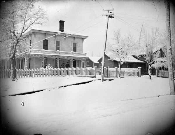 View across street towards two women posing on the porch of two-story wooden house surrounded by snow. The woman on the left is identified as the wife of Charles Van Schaick, and the house is identified as the home of Charles Van Schaick, located at the top of the hill on the west side of Main Street.