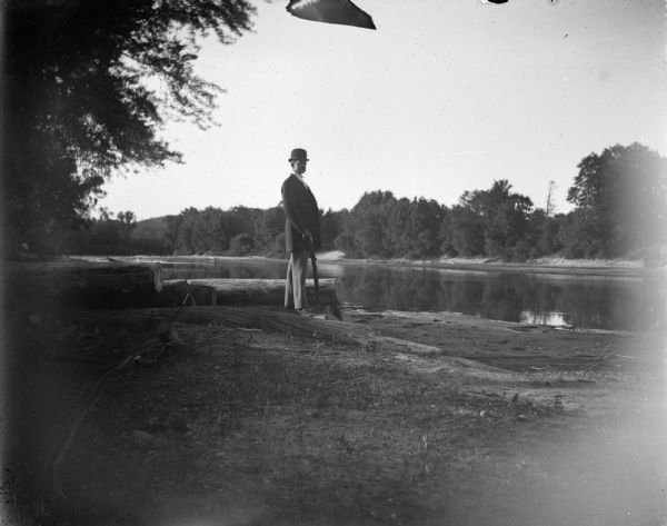 Outdoor portrait of a man posing standing near the water's edge wearing a suit coat and derby hat, and holding an umbrella. Man identified as Charles J. Van Schaick or his brother Tom.