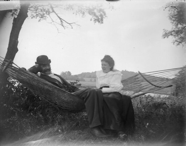 Outdoor portrait of a man and woman posparties in a wedding sitting in a hammock near a body of water. They are identified as Tom Van Schaick and Julia Ormsby.