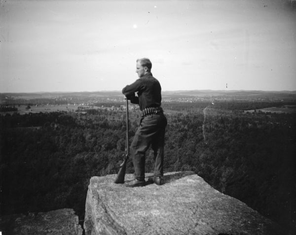 Outdoor portrait of a man posing standing on a rocky precipice and leaning on a shot gun. The man is identified as Roy Van Schaick.
