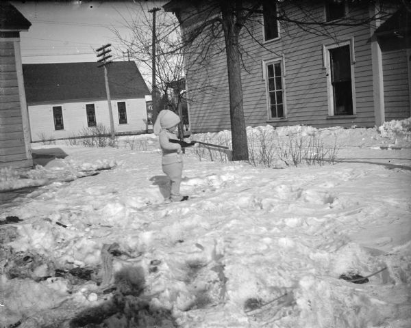 Outdoor portrait of a small child with a toy gun posing standing on snow covered ground by wooden buildings. The house on the right is identified as the home of Charles Van Schaick on the west side of Main Street on top of the hill. The Catholic Church is partially visible across the street.