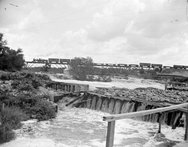 Outdoor view of the dam in front of Black River Falls, showing the rushing water with a wooden railing in the foreground. There is a train on the railroad bridge in the distance. The photograph was taken from the power house located just north of town and shows where the water entered under the power house.