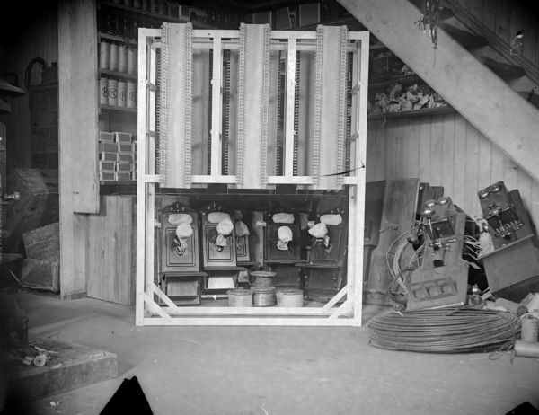Interior view of telephone equipment stored under wooden steps, including receivers and a switchboard. Probably inside the Van Schaick building.
