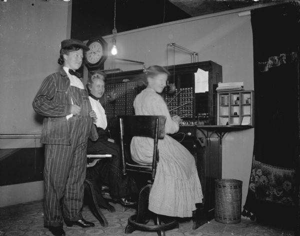 Indoor group portrait of an office space. with two women posing sitting at a telephone switchboard, and another woman posing standing. The woman standing is wearing matching striped overalls, cap, and bow tie. Identified as the telephone exchange office at 13 South First Street.