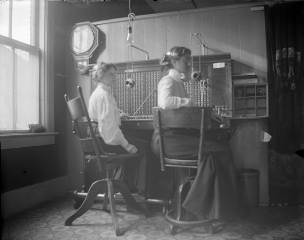 Indoor portrait of an office space with two women posing sitting at a telephone switchboard. Above the switchboard is a clock. There is a window on the left, and a curtain on the right. Identified as probably the telephone exchange office at 13 South First Street.