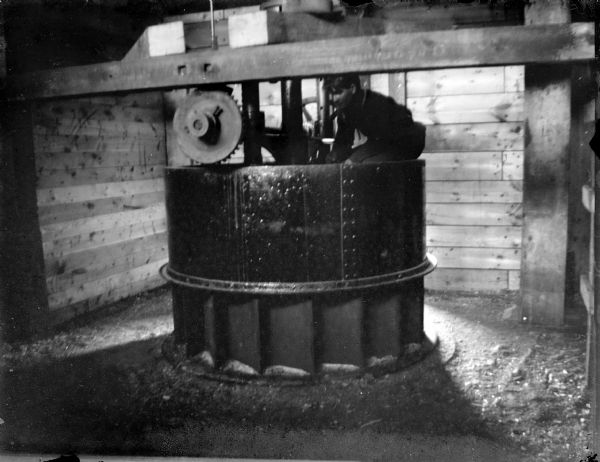 Indoor view of a man posing sitting inside and on top of a piece of large, round machinery. Identified as the interior of the power house.