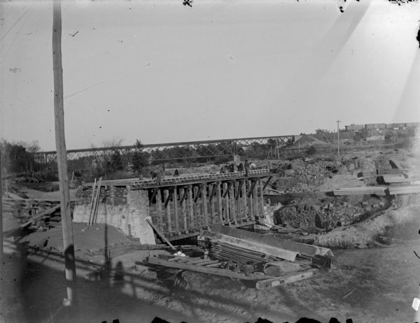Outdoor view looking down at men working on a high brick foundation on the river, with a railroad bridge in the background. Identified as the laying of the foundation for the second powerhouse which was moved north in 1910.