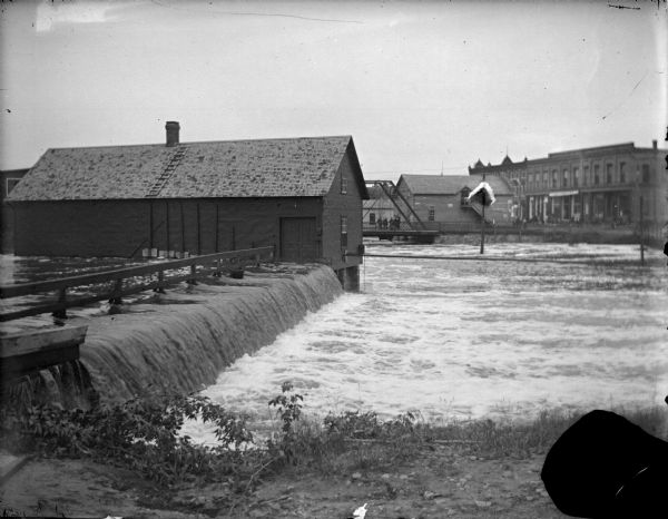 Outdoor view from shoreline towards a powerhouse on the river, with town buildings in the background on the opposite shoreline. Identified as the second powerhouse that was replaced in 1910.