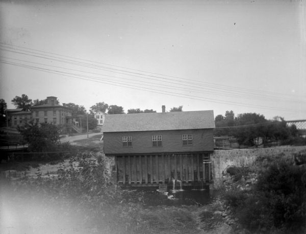 Outdoor view looking down a hill towards a powerhouse on the river, with town buildings on a hill in the background. Identified as the second powerhouse that was replaced in 1910.