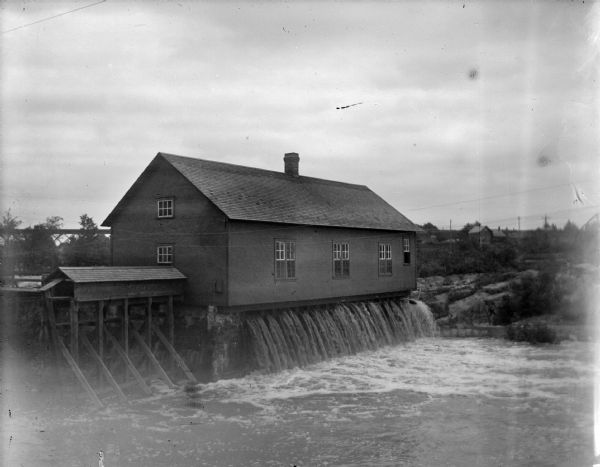 Outdoor view looking down at a powerhouse on the river, with town buildings in the background. Identified as the second powerhouse that was replaced in 1910.