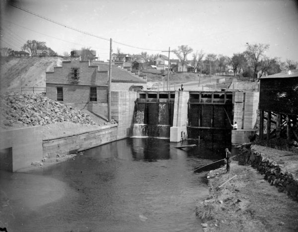Outdoor view looking down at a powerhouse, with town buildings in the background and men fishing along the shoreline. Identified as the third powerhouse built in 1910.