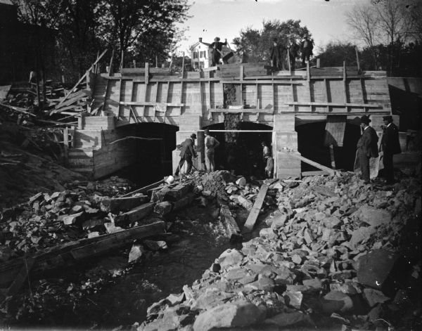 View looking toward men working on a construction site. Men are standing and watching from the rocky shoreline on the right. Identified as the construction of the bulkhead for the powerhouse in 1908.