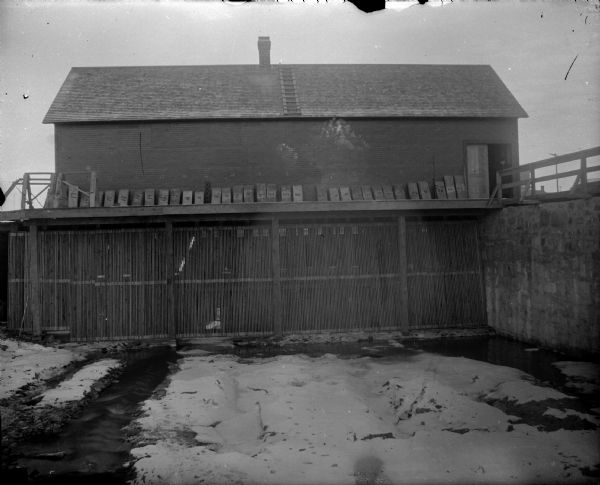 View across snowy yard towards a river powerhouse surrounded by snow. Identified as the second powerhouse.