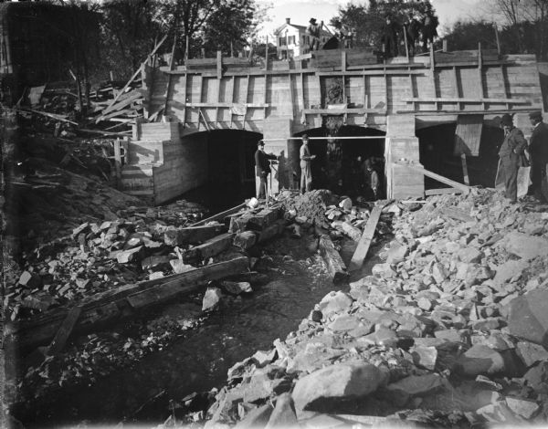 View looking towards men working on a construction site. Identified as the construction of the bulkhead for the powerhouse in 1908. There are men standing and watching on the rocky shoreline on the right.