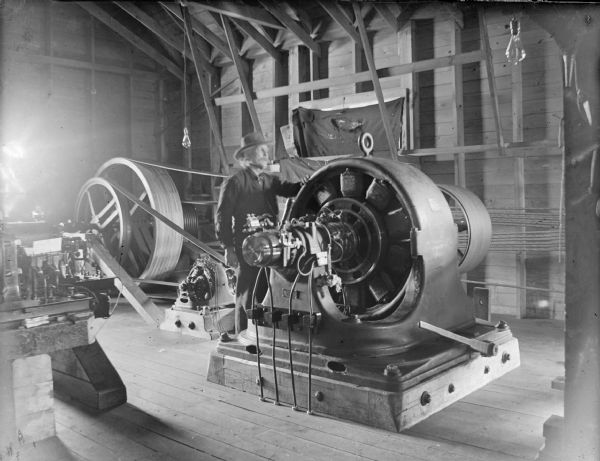 View of a man working near a large piece of belt-driven machinery inside a building. Identified as the interior of a powerhouse.