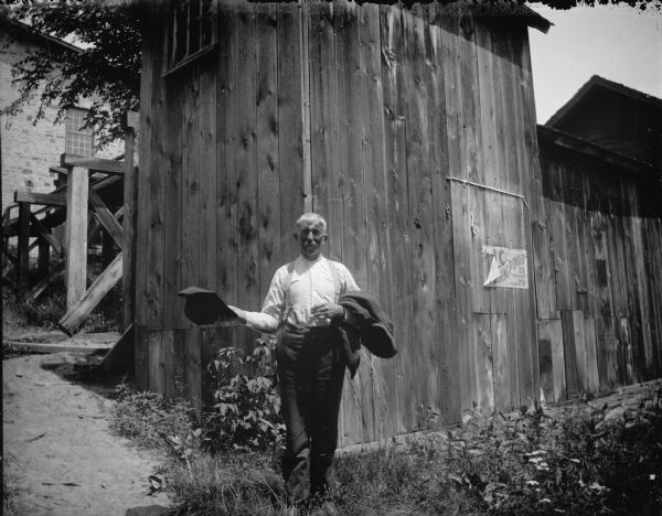 Outdoor portrait of a man posing standing in front of wooden and brick structures. He is holding a hat in his right hand and a coat draped over his left arm. The location is identified as the rear end of the house where power was generated from the powerhouse.