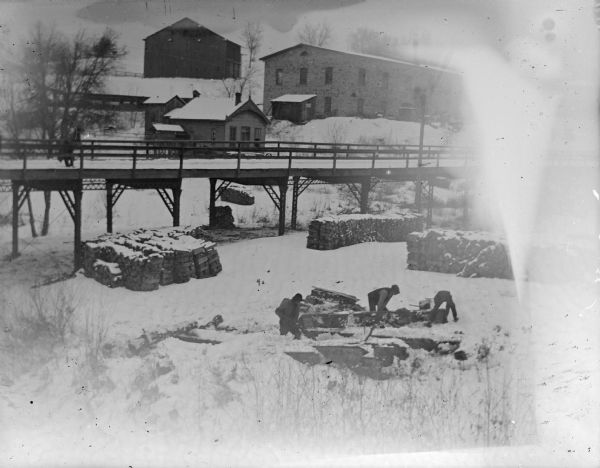 Exterior portrait of men chopping wood on a snow covered-ground with several town structures in the background. The small building in the center was the powerhouse that housed the generator that provided electricity for the town via wires that ran under the bridge. The wood probably came from the Spaulding wagon shop for a cost of one dollar per cord. The view is probably looking northwest from the German Hill Bridge.
