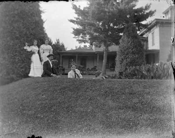 Outdoor group portrait of a man and two women posing sitting on the lawn just behind a low hill, and two women posing standing behind them near tall bushes, in the yard of a large house in the background. Identified as the Spaulding family in the yard of their residence.