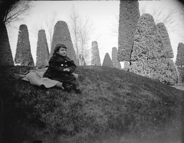 Outdoor portrait of a small girl wearing a winter coat posing sitting on the lawn of a well-manicured yard with sculpted shrubs. Location identified as the Spaulding residence.