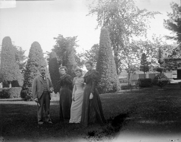 Outdoor group portrait of a man and three women posing standing on the lawn of a well-manicured yard with sculpted shrubs. Identified as the Spaulding family in the yard of their residence.