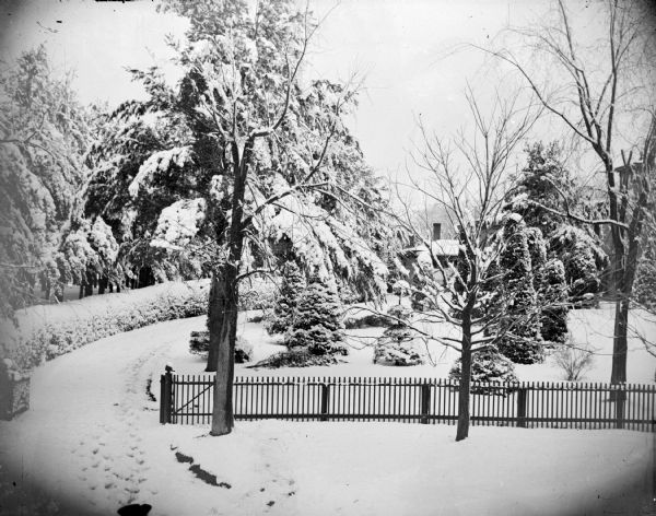 Elevated view of fence and drive, with a large house behind trees and shrubs in a snow-covered yard. Identified as the Spaulding residence.