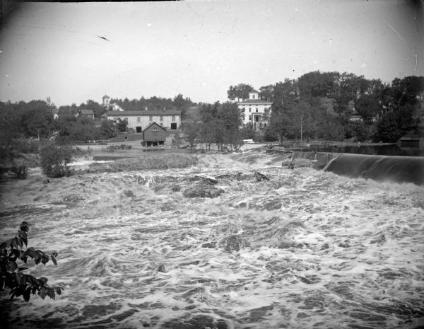 View across river just below the dam towards buildings on the opposite shoreline. The buildings identified include the Spaulding wagon shop on the left, and the Spaulding boarding house on the right.