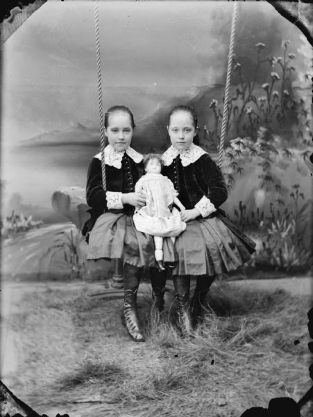Studio portrait in front of a painted backdrop of twin European American girls posing sitting on a swing and holding a doll on their laps between them. Identified as Jane and Mary Spaulding.