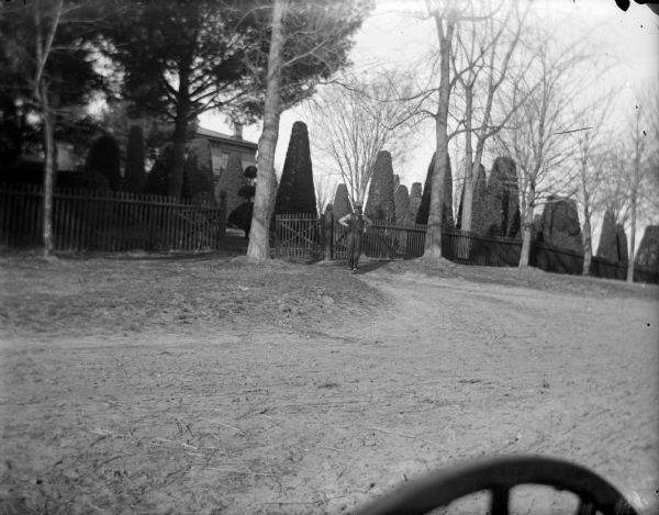 View across unpaved street towards a man posing standing near the front of a picket fence. Behind the fence is a well-manicured yard with sculpted shrubs. Identified as the yard of the Spaulding residence.