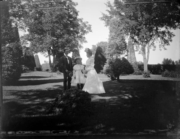 Outdoor group portrait of a man, woman and girl posing standing in a well-manicured yard with sculpted shrubs. Identified as the yard of the Spaulding residence.